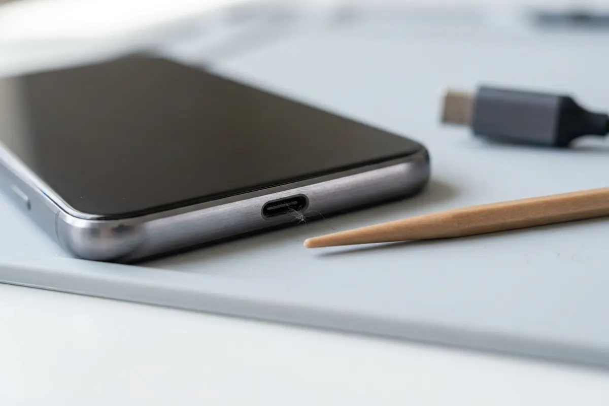 Makroaufnahme einer leeren USB-C-Buchse am unteren Rand eines Smartphones, daneben ein Holzstäbchen zur Reinigung